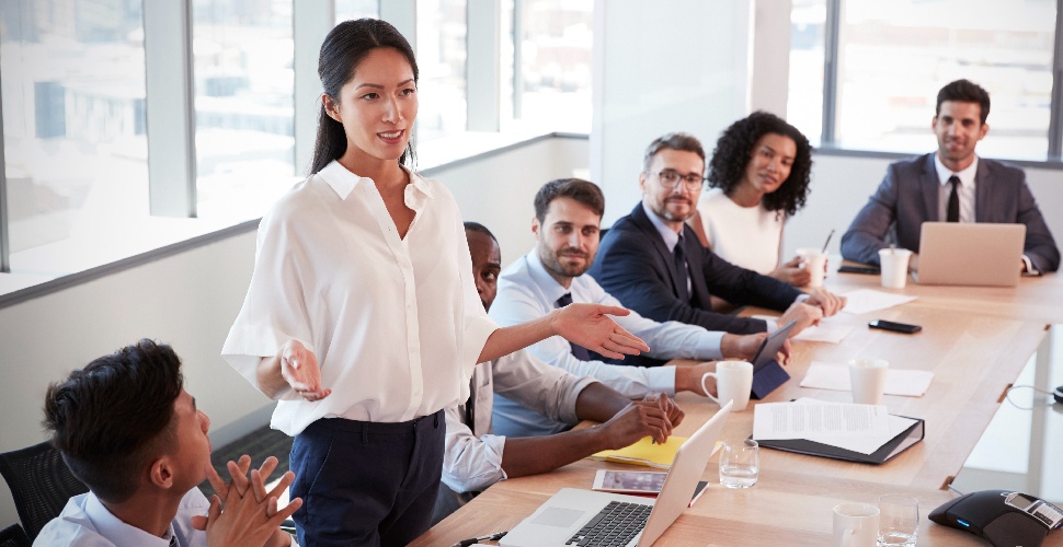 A group of white-collar professionals having a meeting, with one Asian female standing and speaking