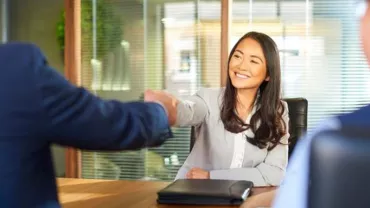 A woman smiles while shaking hands with a person across a table in an office setting.