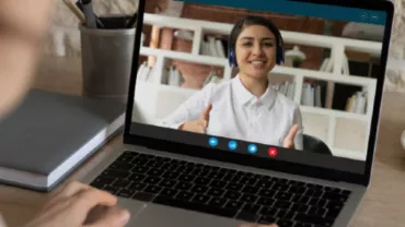 Person video calling on laptop at desk with coffee and notebooks.