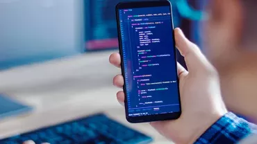 Person holding a smartphone displaying code, with a keyboard and monitor in the background.