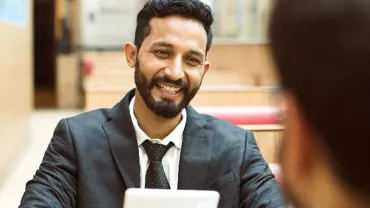 Smiling man in a suit holding a tablet, sitting at a table.