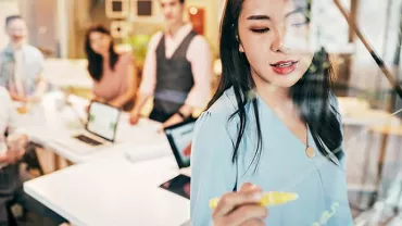 Woman writing on a glass board during a team meeting in an office.