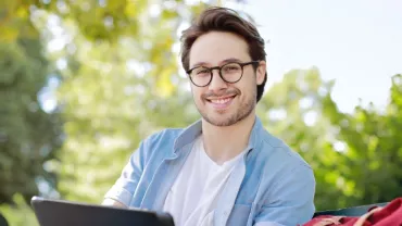 Man with glasses smiling outdoors, sitting on a bench.