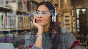 Woman with headphones studies at a library, gazing pensively outside.