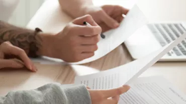 Two people reviewing documents at a desk with a laptop open.