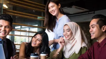 Group of five people smiling at a laptop in a modern office.