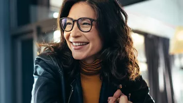 A woman in glasses smiling, sitting indoors with natural light.