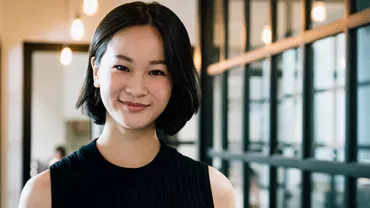 A smiling person stands in a modern hallway with glass walls.