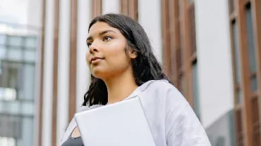 Person holding a laptop outdoors, standing in front of a modern building.