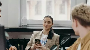 Three people in an office, one holding a phone, engaged in conversation.