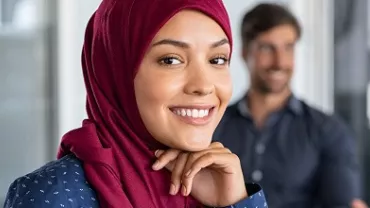 A female professional smiling, on a office background with three people dressed in corporate wear seated behind her