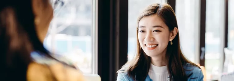 Two women sitting at a table, one smiling, in a bright, window-lit setting.